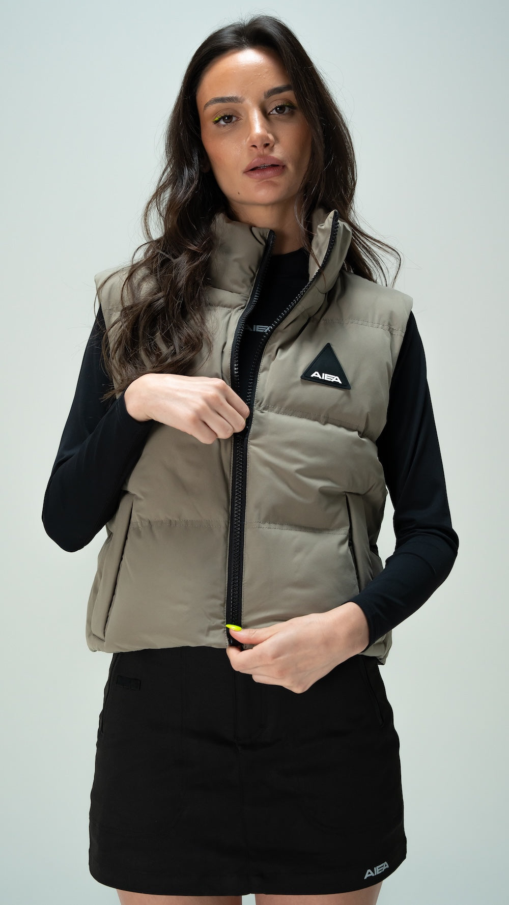 A woman modeling a light taupe puffer vest with a black zipper and a black triangular silicone logo patch on the chest, worn over a black long-sleeve top and a black skirt.