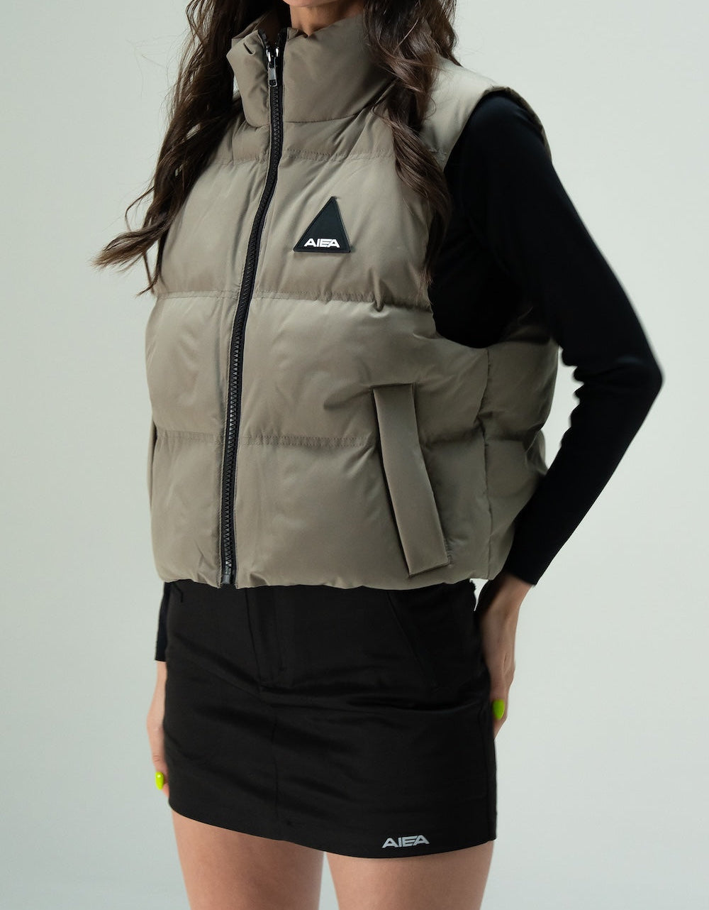 AIEA Cropped Puffer Vest — Woman wearing khaki green puffer vest with a black triangular silicone logo patch on the chest, worn over a black long-sleeve top and a black skirt.